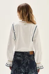 Blusa blanca de algodón con cuerpo de jersey y mangas de popelina. Presenta detalles de punto smock en puños y cuello, adornados con delicados bordados en azul y dorado.
