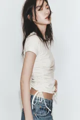 Camiseta corta de cuello redondo y manga corta, color gris oscuro, con frunces laterales ajustables mediante cordones.