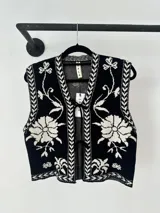 Chaleco tejido de punto con diseño floral en contraste blanco y negro. Presenta cierre frontal mediante lazos y bordes con detalle geométrico.