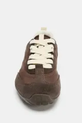 Zapatilla deportiva de mujer, estilo running, color marrón oscuro con detalles en color crema/beige claro en el talón y cordones. Presenta una combinación de materiales, incluyendo gamuza en puntera y ojales, y tejido sintético en el cuerpo.