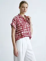 Vestido camisero de silueta relajada, con cuello clásico, cierre frontal de botones y mangas cortas con dobladillo. Presenta un estampado de cuadrícula irregular en color rojo sobre fondo blanco.