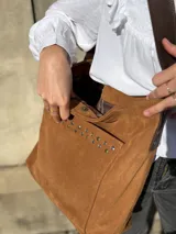 Cartera tipo tote bag de cuero color camel, con detalles de tachas metálicas en el bolsillo frontal y tiras largas para el hombro.