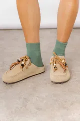 Zueco de cuero gamuzado color tostado, con cordones decorativos adornados con cuentas de madera y piezas envueltas en cuero. Diseño tipo mocasín con costuras visibles en la parte superior y abierto en la parte trasera.