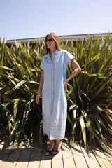 Vestido camisero de lino color lila con bordados rojos en contraste. Presenta un diseño de largo midi con cuello camisero, cierre frontal de botones y mangas cortas. Los detalles bordados recorren la botonadura, los puños y el ruedo de la prenda.