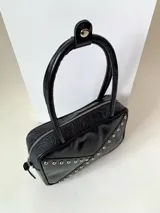 Cartera rígida negra con detalles en rojo y tachas plateadas. Tiene herrajes y cierre metálico plateado marca YKK, asa fina para llevar al hombro o en la mano y correa larga extra regulable. El interior es forrado, tiene un bolsillo con cierre enmarcado en cuero, y un segundo bolsillo plaqué.