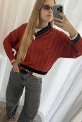 Buzo tejido de punto con escote en V, manga larga y diseño holgado. Presenta un patrón de rayas verticales en relieve y detalles de franjas en contraste en el cuello, puños y ruedo. Incluye un pequeño bordado en el pecho.