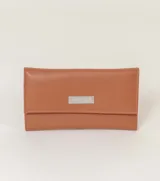 Billetera tipo sobre color champagne, con textura de cuero granulado y cierre de solapa. Presenta una placa metálica rectangular con el nombre de la marca grabado en el frente.