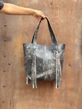 Bolso tipo tote color beige con textura perforada, asas largas y detalles de flecos largos en los laterales. Cuenta con un bolsillo frontal exterior.