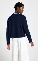 Cardigan de punto azul marino con cuello polo, cierre frontal de botones y detalle de bordado en el pecho. El cuello presenta un borde festoneado en color blanco.