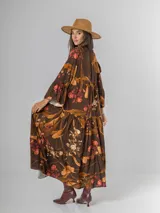Vestido largo de manga larga, corte oversized y cuello camisero con abertura en V y botones. Presenta un estampado botánico en tonos tierra sobre fondo marrón oscuro, con volados en el ruedo.