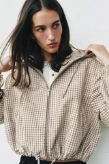 Campera estilo cazadora con capucha ajustable con cordones, cierre frontal de cremallera, mangas largas con puños elásticos, bolsillos delanteros de doble vivo y bajo ajustable con cordones. Presenta un diseño de cuadros en tonos beige y marrón.