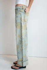 Jeans de tiro bajo confeccionados en denim, con diseño holgado y estampado integral de rosas en tonos amarillos y verdes sobre fondo celeste claro. Cuenta con cinco bolsillos, trabillas para cinto y botamangas con vuelta.