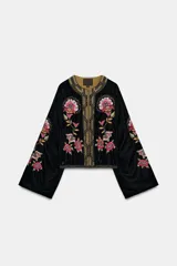 Chaqueta abierta de terciopelo negro con cuello redondo y mangas largas acampanadas. Presenta bordados florales en tonos rosados y verdes, con aplicaciones de abalorios y detalles dorados en el cuello y la parte frontal.