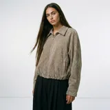 Chaqueta de textura aterciopelada en color beige, con cuello clásico, cierre frontal completo y puños con botones. Presenta un diseño de corte recto con bolsillos laterales.