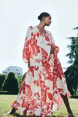 Vestido largo de tejido ligero color blanco con estampado floral rojo. Tiene cuello redondo con lazos, mangas globo largas con puños abotonados, corte holgado y falda con volantes en el ruedo.