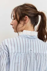 Camisa de rayas celestes y marrones, de corte holgado y manga larga, con cuello en V y detalles blancos en el cuello y los puños.