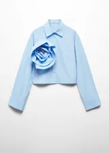 Camisa crop de color celeste con cuello camisero, manga larga con puños abotonados y cierre frontal de botones. Destaca por una maxi flor decorativa aplicada en el pecho.