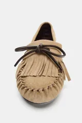 Mocasín plano de gamuza color tostado, con detalle de flecos y lazo de cuero marrón en la parte delantera. Acabado en punta redonda y suela de 1 cm.