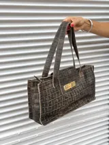 Cartera tipo tote bag de diseño rectangular, confeccionada en cuero negro con acabado brillante y efecto arrugado. Presenta doble asa de mano, cierre frontal con solapa y herraje metálico dorado tipo torniquete.