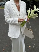 Blazer de corte sastre en color blanco, confeccionado en crepé satén con detalles de flecos de seda en toda la prenda. Presenta solapas clásicas, cierre frontal con botón de nácar y dos bolsillos laterales tipo ojal.