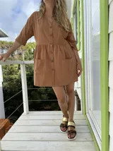 Vestido corto color marrón, con cuello en V, mangas 3/4, cierre frontal con botones, dos bolsillos frontales y corte a la cintura con falda fruncida.