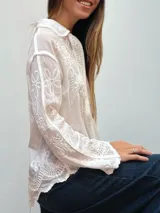 Camisa de manga larga en color blanco, confeccionada en tela semitransparente con detalles de bordados florales y geométricos en todo el diseño. Presenta cuello clásico, cierre frontal con botones y terminaciones con bordes festoneados.