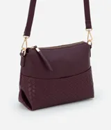 Bolso bandolera pequeño color borgoña, confeccionado en poliuretano con detalle texturizado tejido en la parte inferior. Cuenta con correa transversal ajustable, cierre de cremallera y bolsillo frontal con botón.