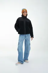 Campera negra de estilo urbano, confeccionada en una combinación de tela técnica y textura tipo corderito (fleece). Presenta cierre frontal con solapa y botones a presión, cuello alto y logo estampado en el pecho.