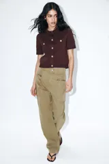 Chaqueta de punto color marrón oscuro, con cuello redondo, manga corta y cierre frontal de botones dorados. Presenta dos bolsillos falsos con botón dorado en la parte delantera superior.