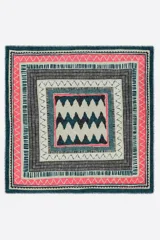 Chal cuadrado con estampado geométrico en capas concéntricas. Predominan los colores rosa coral, verde azulado oscuro, gris oscuro y beige claro. Presenta patrones de zigzag y líneas verticales.
