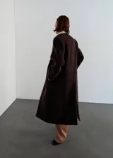 Abrigo largo de lana color marrón oscuro, con diseño recto, cuello en pico sin solapas, manga larga, bolsillos laterales y cierre frontal de un botón con detalles metálicos dorados.