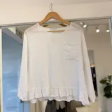 Blusa blanca de lino, con cuello redondo, mangas largas y un bolsillo frontal aplicado con detalles bordados. Cuenta con un volante fruncido en el borde inferior.