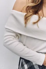 Buzo de punto color blanco, con escote tipo bote que deja los hombros al descubierto y mangas largas ajustadas.