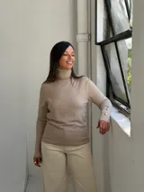 Polera de cuello alto en color visón, confeccionada en tejido de punto fino. Presenta mangas largas con puños acanalados decorados con tres botones dorados en cada uno.