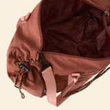 Bolso de deporte color marrón con detalles en rosa claro. Presenta correas de mano tipo cuerda, una correa ajustable para el hombro, un sistema de cordones elásticos decorativos en el frente y bolsillos laterales con cierre.