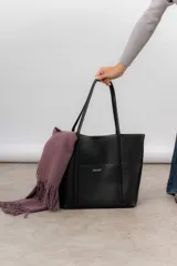 Bolso tipo tote de color marrón, confeccionado en material símil cuero texturado. Presenta un diseño estructurado con asas largas para llevar al hombro y el logo de la marca aplicado en la parte frontal.