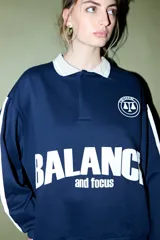 Buzo azul marino con cuello polo blanco y cierre parcial. Presenta un estampado frontal con la palabra "BALANCE" en blanco y "and focus" debajo, junto con un logo circular en el pecho. Tiene rayas blancas en las mangas.
