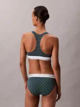 Bralette deportivo sin relleno ni forro, con diseño de espalda cruzada y banda elástica inferior con el logo de la marca. Presenta un estampado geométrico con el monograma de Calvin Klein en tonos verde oscuro y negro.