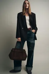 Bolso tipo citybag confeccionado en serraje color marrón oscuro. Presenta una solapa frontal con cierre magnético, asas de hombro integradas y detalles de hebillas metálicas en los laterales para ajuste. Incluye bolsillo interior con cremallera.