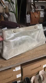 Sobre de mano tipo clutch confeccionado en cuero con acabado metalizado plateado. Presenta un diseño rectangular con cierre superior mediante solapa y dos botones a presión metálicos.