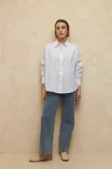 Camisa de corte clásico y holgado, confeccionada en tela blanca, con cuello camisero, cierre frontal de botones de nácar y mangas largas con puños abotonados.