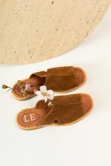 Sandalias planas de gamuza color marrón con diseño de tira ancha y aberturas laterales. Presentan una plantilla de cuero con el logo de la marca grabado.