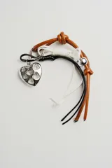 Llavero con dije metálico plateado en forma de corazón calado, cordones de cuero marrón y sintéticos en blanco y negro, y cascabel plateado.