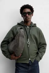 Bolso de hombro tipo media luna, fabricado en piel craquelada color marrón oscuro. Tiene un compartimento principal con cierre de cremallera y un bolsillo interior también con cremallera.