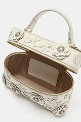 Bolso de mano formato vanity color crudo, con apliques de flores en relieve y hojas en el cuerpo. Cuenta con asa corta superior y cierre con cremallera doble dorada.