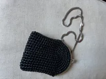 Cartera de mano tejida a crochet color negro, con cierre metálico plateado y cadena larga delgada. Presenta aplique de flor con strass en el frente.