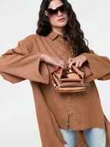 Camisa larga de lino color marrón, con corte oversize, mangas anchas y bolsillo frontal de cuero sintético en tono contrastante. Presenta cierre frontal con botones y diseño de bajo irregular.