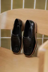 Mocasines negros de cuero con textura tipo cocodrilo en la parte superior y acabado brillante. Presentan una banda frontal clásica y una suela plana con detalle de borde en color natural.