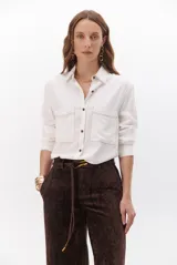 Camisa beige de manga larga, con cuello clásico, botones marrones y dos bolsillos frontales. Presenta costuras decorativas en contraste.