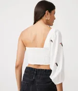 Blusa cropped de lino blanca con manga tres cuartos, hombro asimétrico, escote corazón, lastex en la espalda y bordados de tucanes.